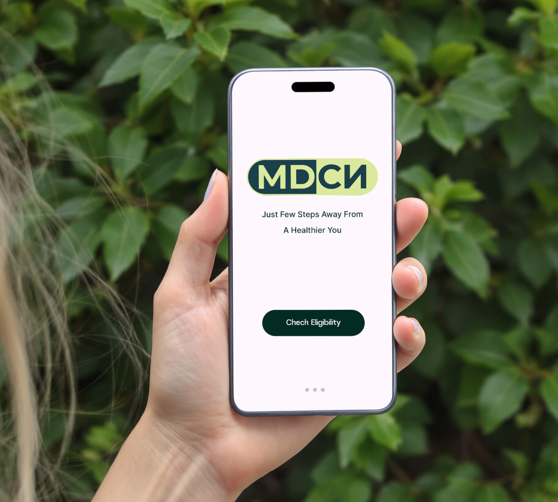 A hand holds a smartphone displaying an app titled "MDCN" with the text "Just Few Steps Away From A Healthier You" and a button labeled "Check Eligibility," against a leafy background.
