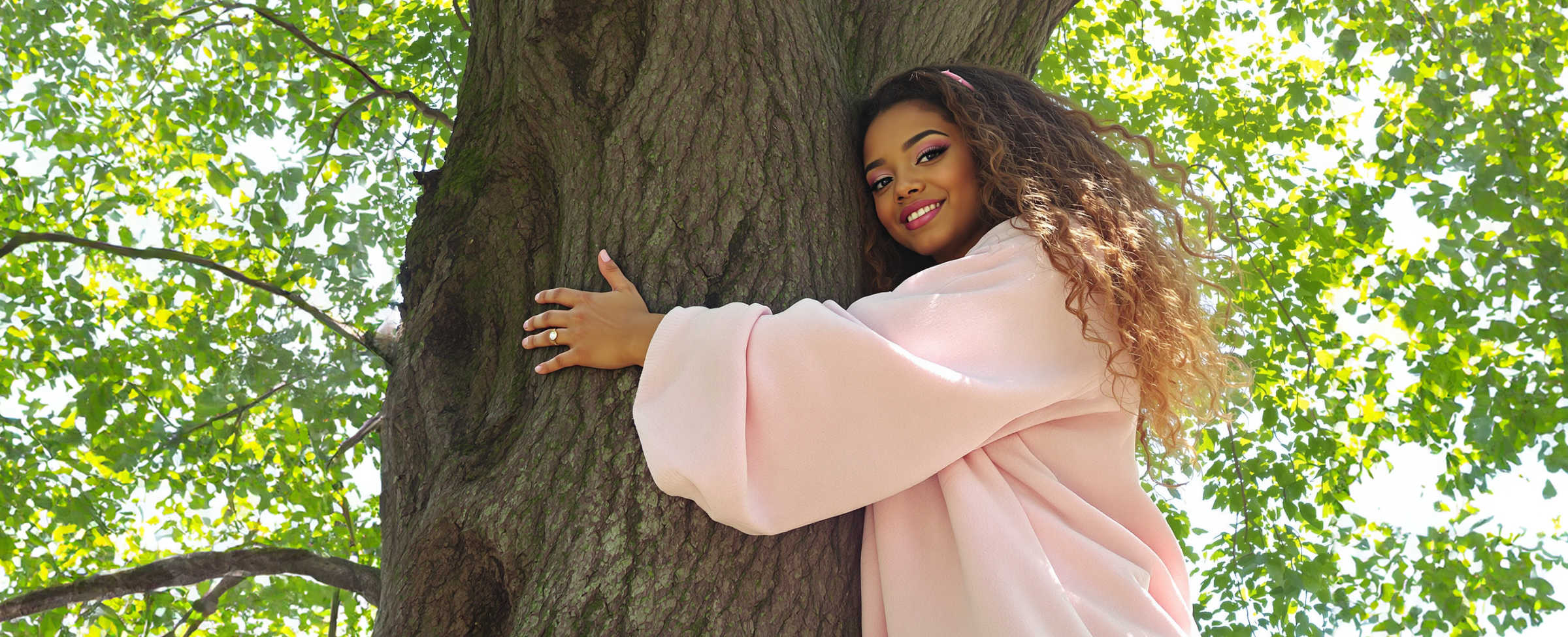 Person in a pink coat hugging a large tree with a cheerful expression, surrounded by green leaves.
